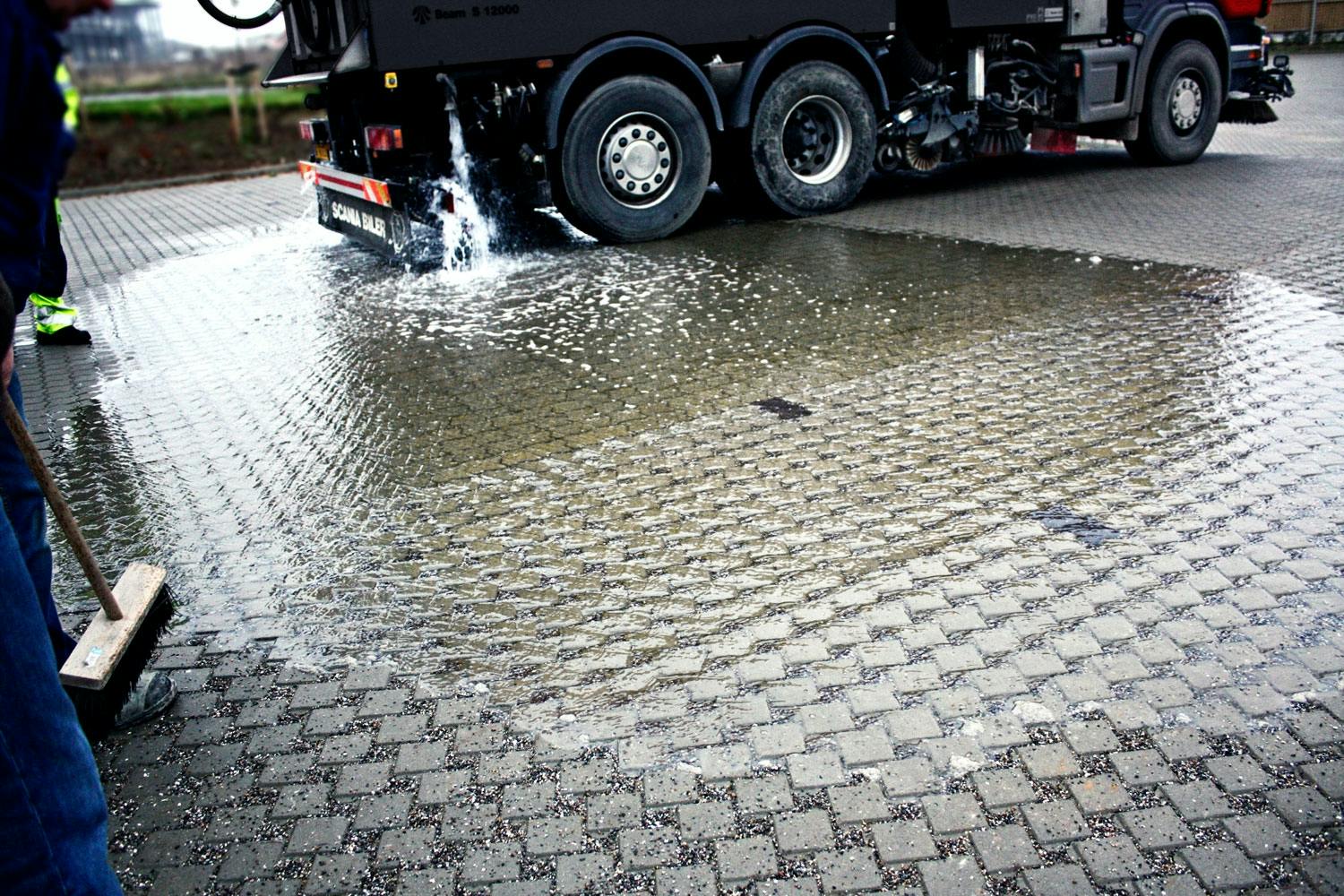 Ved å tømme en tank full av vann over steinene, demonstreres det her hvor effektivt den drenerende belegningen suger til seg vannet. Et bare 15 cm tykt drenerende lag kan uten problemer romme et voldsomt skybrudd med 50 mm nedbør. Fra grusen siver vannet senere ned i bakken, som normalt.